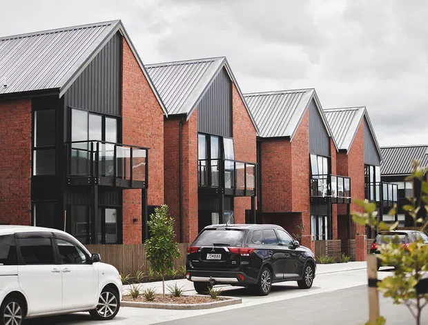 Exterior view of a contemporary multi-unit residential development with repeated narrow gabled forms, red brick walls, dark vertical cladding, silver metal pitched roofs, black-framed glazing, and small balconies. Cars and landscaping are present in the foreground, with an overcast sky and neighbouring buildings in the background.