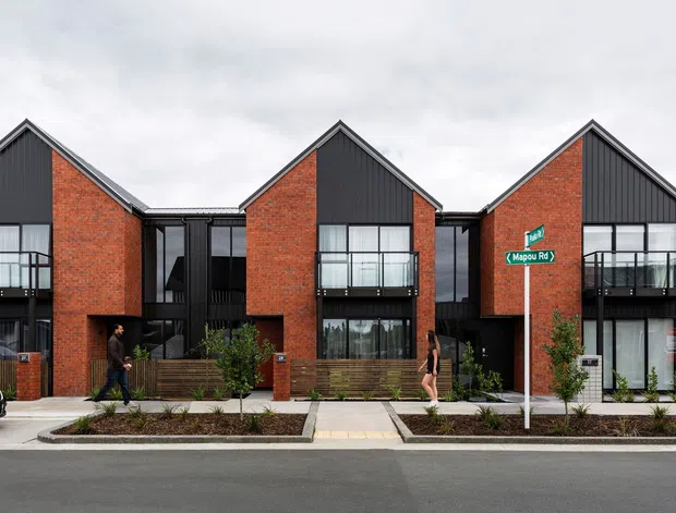 Street-facing view of a contemporary multi-unit residential development with three prominent gabled townhouse facades. The buildings combine red brick walls, dark vertical cladding, extensive black-framed glazing, balconies, and recessed entrances. A landscaped sidewalk and street signs are visible in the foreground.