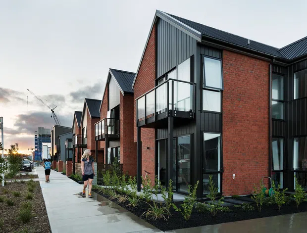 Street-level exterior view of a contemporary multi-unit residential development with repeated steep gabled roof forms, red brick facades, dark vertical metal cladding, black-framed glazing, and small glass-balustraded balconies. A landscaped pedestrian frontage and concrete footpath run alongside the row of homes.