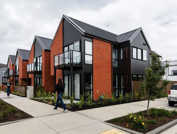 Exterior view of a contemporary multi-unit residential development with repeated steep gabled roof forms, red brick walls, black vertical cladding, large black-framed windows, and projecting balconies. Concrete pedestrian paths, landscaped planting beds, and neighbouring townhouses are visible in the foreground and background.
