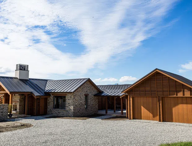 Exterior view of a low-rise residential property arranged around a broad gravel forecourt. The building combines dark grey metal gabled roofs, natural irregular stone masonry walls, and warm timber-clad gabled garage or outbuilding forms. A covered link connects the separate volumes, with a chimney visible on the left. Bright blue sky and open landscaping surround the house.