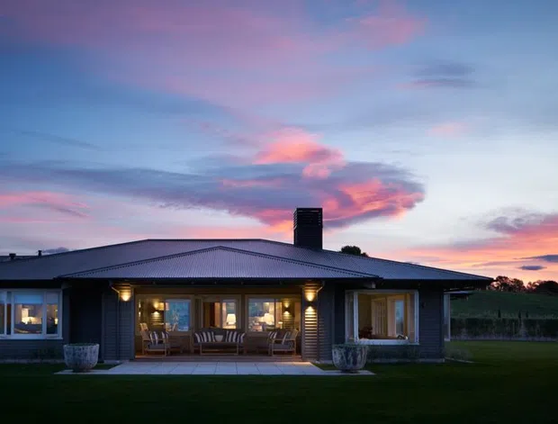 Single-storey residential house photographed at dusk, with a broad low-pitched roof, dark horizontal cladding, a central chimney, and a covered veranda with warm lighting. Large windows and glazed doors open toward a neatly landscaped lawn, with rural vegetation visible beyond and a colourful sunset sky.