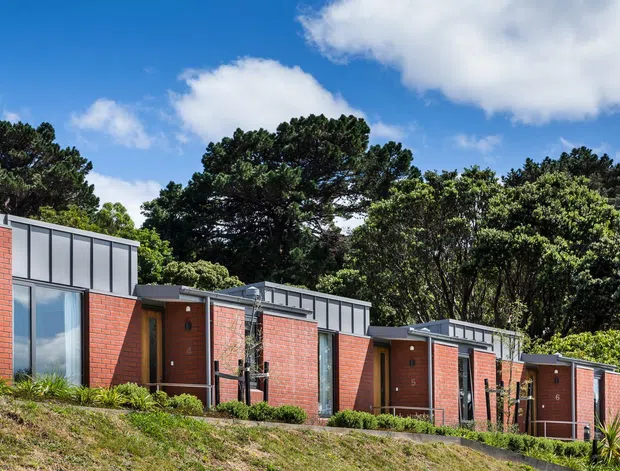 Exterior view of a low-rise contemporary brick development composed of several compact attached or closely grouped units. The buildings feature red brick walls, dark metal-framed clerestory-style parapet panels, simple flat or low-pitched roof forms, and tall glazed openings with timber or dark doors. The complex sits on a landscaped sloping site bordered by mature trees under a bright blue sky.