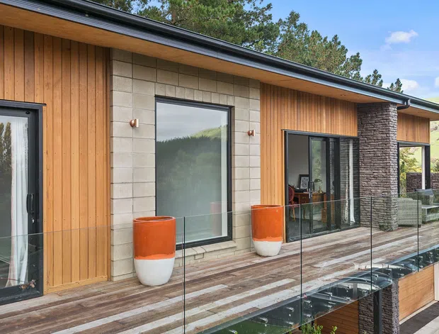 Contemporary single-storey building exterior with a long covered terrace and frameless glass balustrade. The facade combines vertical timber cladding, pale masonry blockwork, dark stacked-stone feature walls, extensive dark-framed glazing, and a low-pitched roof. Bright orange-and-white planters add colour along the timber deck, with trees and a green hillside visible beyond.