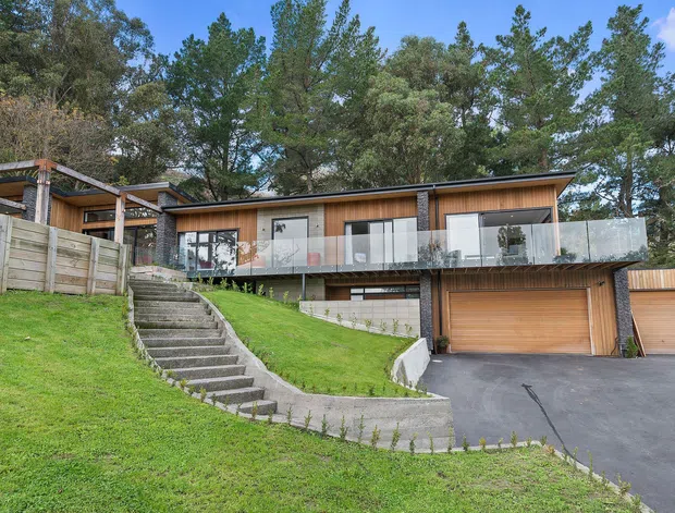 Exterior view of a contemporary, split-level timber-clad house on a sloped, landscaped site. The home features a long low-pitched roofline, large glazed openings, a glass-balustraded upper terrace, exposed structural posts, and an integrated double garage accessed from a paved driveway. Concrete steps and retaining walls lead through the lawn to the main level, with mature trees surrounding the property.