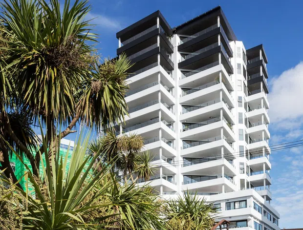 Exterior view of a tall modern residential apartment building with stacked balconies, white facade, dark roof and balcony bands, and extensive glazing. The building is framed by landscaped plantings and a timber stairway with metal handrails in the foreground under a bright blue sky.