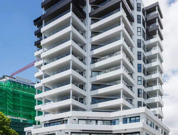 Street-level exterior photograph of a tall contemporary apartment building with a stepped, angular massing and numerous projecting balconies. The facade is predominantly white with dark charcoal balcony soffits, railings, and upper-storey cladding, along with extensive glazing. A ground-level entry and parking area are visible, with an adjacent building under construction. Bright blue sky and urban streetscape frame the building.