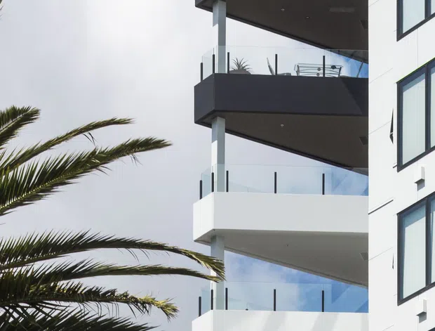 Close-up exterior view of a contemporary multi-storey building with stacked projecting balconies, clear glass balustrades, dark soffits and balcony fascias, and a white facade with dark-framed windows. Palm fronds occupy the foreground against a bright blue sky, suggesting a warm urban or coastal setting.