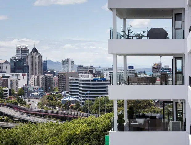 Exterior view of a contemporary multi-storey apartment building with stacked white balconies, clear glass balustrades, dark-framed glazing, and outdoor furniture. The building overlooks a dense urban skyline with transport infrastructure and distant hills.