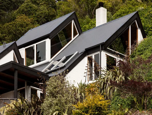 Exterior view of a contemporary multi-level home set within dense, established landscaping. The house has sharply pitched dark metal gable roofs, white rendered or painted walls, extensive dark-framed glazing, projecting roof forms, and a prominent white chimney. A covered outdoor structure is visible at the lower left, while layered planting partially screens the facade.