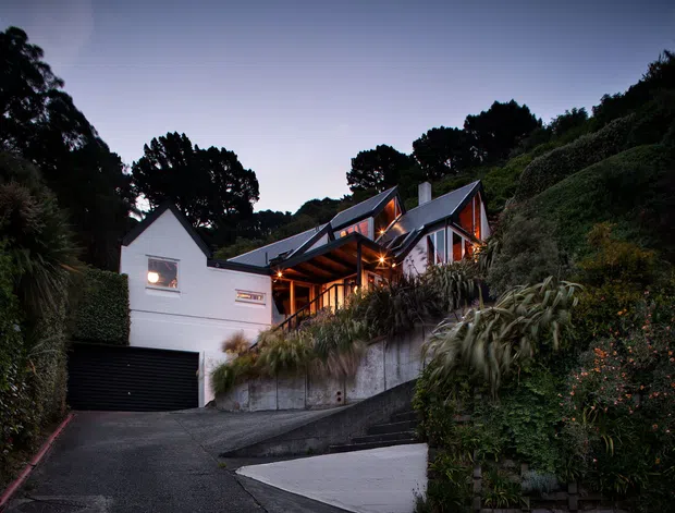 Twilight exterior view of a contemporary multi-level hillside home, with white rendered walls, steep intersecting pitched roofs, dark roof cladding, extensive glazing, warm timber-lined covered entry, and an integrated black garage. The house is set into a densely planted sloping landscape with retaining walls and a steep driveway.