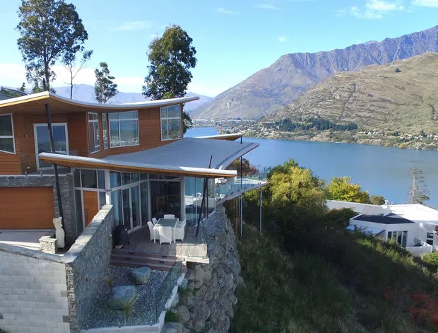 Exterior view of a contemporary multi-level hillside house with warm timber cladding, extensive glazing, broad roof overhangs, and stone retaining walls. The home overlooks a lake and rugged mountain landscape, with a covered outdoor dining terrace visible below.