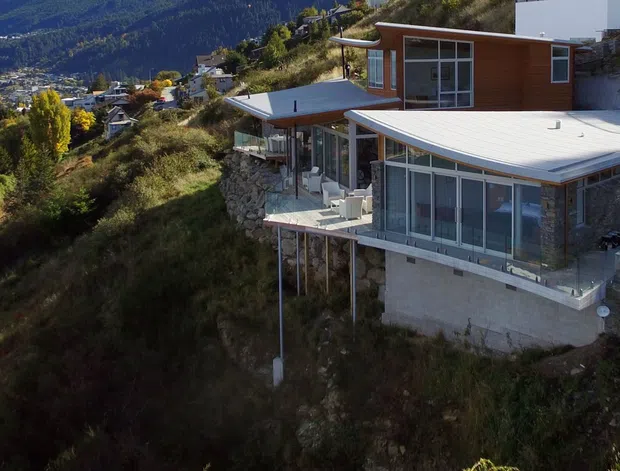 A contemporary multi-level hillside house viewed from outside, perched on a steep landscaped site with a broad white low-pitched roof, extensive floor-to-ceiling glazing, timber-clad upper walls, concrete lower structure, terraces, and slender structural supports. A dense residential landscape and mountain valley are visible in the background.