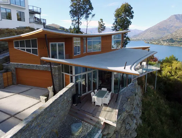Exterior view of a contemporary multi-level hillside residence overlooking a lake and mountainous landscape. The home combines warm timber wall cladding, extensive glazing, a curved metal roof, stone retaining walls, terraces, and an integrated garage. A neighbouring modern dwelling is partially visible above.