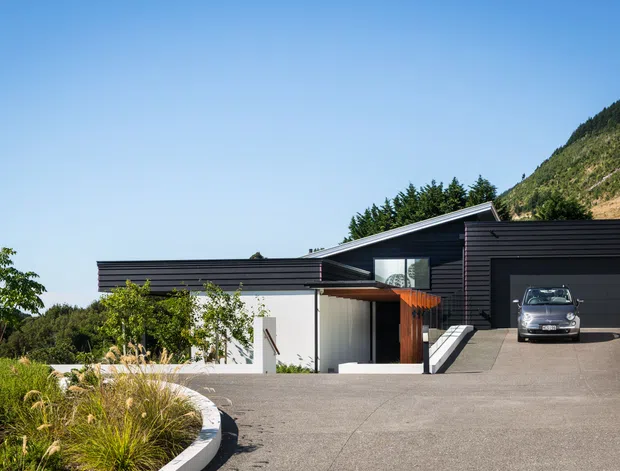 A contemporary single-storey house set into a sloping, wooded site. The home has a low, angular roofline, dark horizontal exterior cladding, white rendered walls, large glazed openings, and an integrated dark garage. A broad concrete driveway and curved landscaped garden edge occupy the foreground, with a steep vegetated hillside behind.