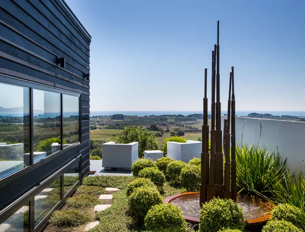 Exterior view of a contemporary dark-clad building overlooking a coastal rural landscape and water. The scene includes expansive glazing, a landscaped terrace with clipped shrubs, ornamental grasses, stepping stones, sculptural vertical timber poles, and a shallow circular water feature. The architecture has clean horizontal lines and a restrained modern composition.