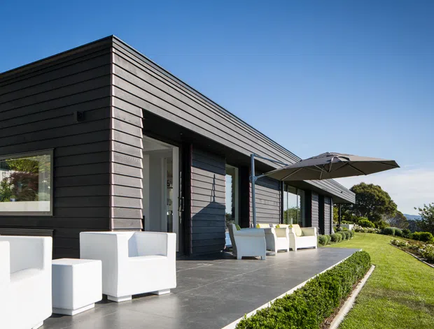 A modern single-storey residence with a low, flat-roofed form and dark horizontal timber-like cladding. Large glazed openings connect the interior to a broad paved terrace furnished with white outdoor seating and a large umbrella. The landscaped lawn slopes toward a distant coastal or water view, with clear blue sky and bright daylight.