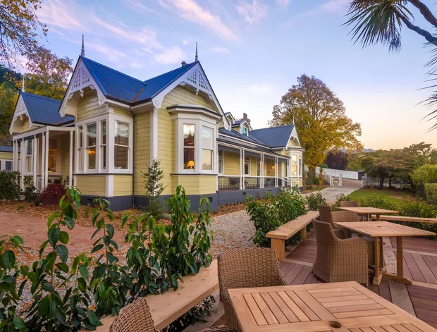 Exterior view of a restored ornate timber house with pale yellow weatherboard cladding, dark blue-grey gabled roof, decorative white bargeboards, bay windows and a long covered veranda. The house sits within landscaped gardens beside a timber outdoor dining deck furnished with tables and wicker chairs.