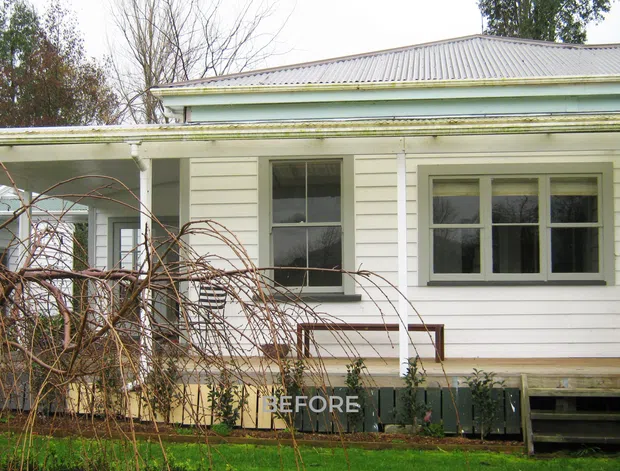 Exterior view of a modest single-storey weatherboard house, showing a covered front veranda, white horizontal cladding, timber-framed windows, a corrugated metal roof, and steps leading to the entry. Leafless garden vegetation partially obscures the facade; a faint “BEFORE” overlay indicates a renovation comparison image.