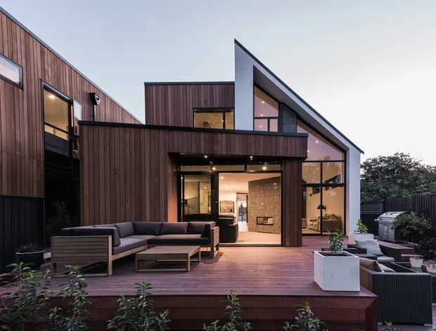Contemporary multi-level residence viewed from a landscaped timber deck and outdoor seating area. The home features dark vertical timber cladding, black-framed glazing, strong angular roof forms, and expansive glazed openings connecting the interior to the deck. Warm interior lighting is visible through the windows and doors.