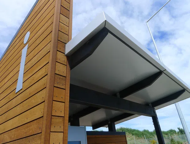 Close-up exterior view of a contemporary building with warm horizontal timber cladding, a projecting flat canopy or roof structure supported by dark steel beams, and a slender vertical white architectural detail. The image is photographed from below against a partly cloudy sky.