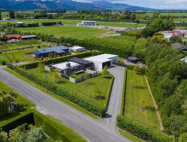 Aerial view of a contemporary residential property in a spacious semi-rural setting. The home has low-profile modern forms, dark and light flat-roofed volumes, extensive glazing, landscaped gardens, paved driveways and neatly maintained lawns. Surrounding farmland, sports fields, hedges and distant mountain ranges are visible.