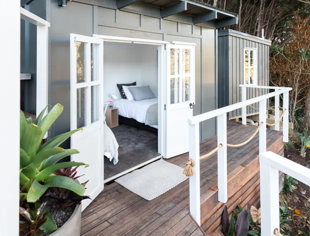 Exterior view of a compact modern cabin or studio with dark vertical cladding, white-framed French doors, and a timber deck with white balustrades and rope detailing. The open doors reveal a simply furnished bedroom with a white bed and grey carpet. Dense planting surrounds the raised deck.