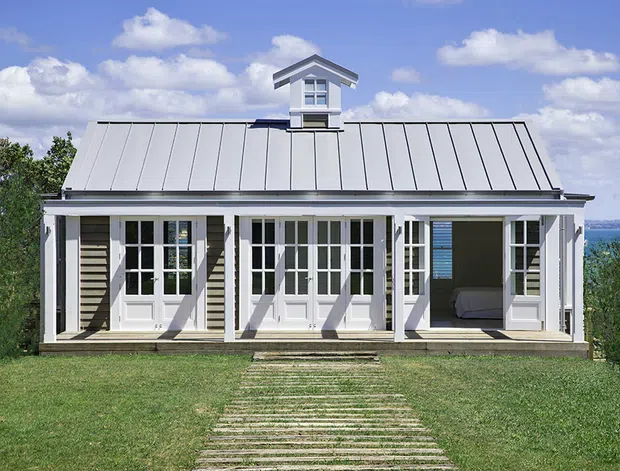 A compact, symmetrical coastal cottage viewed from the front, with a light weatherboard facade, broad covered veranda, repeated white French doors, and a standing-seam metal gable roof with a small central dormer. A grass lawn and simple stepping-stone path lead to the house, with water visible beyond the right side.