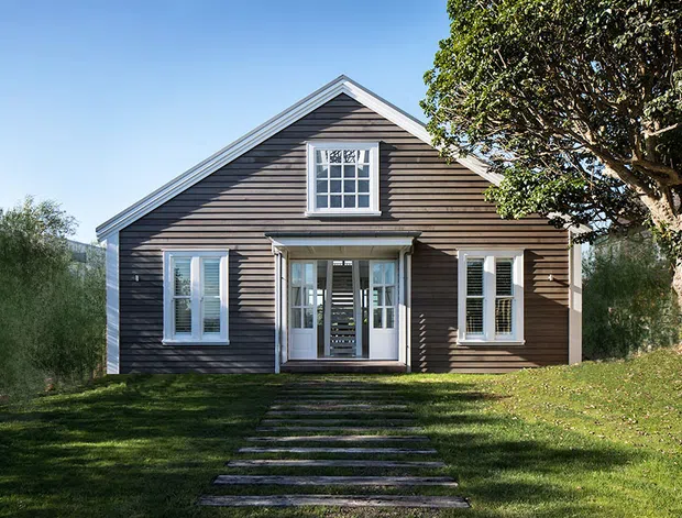 Front exterior view of a compact single-storey gable-roof house with dark horizontal weatherboard cladding, white trim, symmetrical multi-pane windows, and a glazed central entry porch. The house sits in a landscaped grassy setting with stepping-stone access and mature trees.
