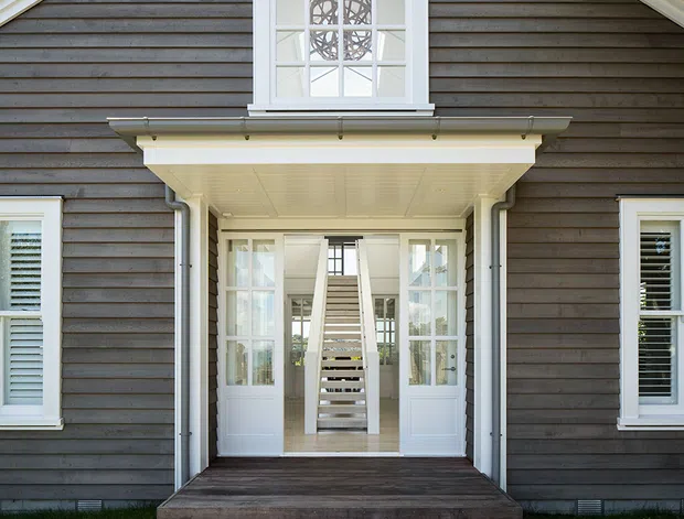 Symmetrical exterior view of a dark weatherboard house entry, featuring a covered porch with white trim, glazed double doors, sidelights, and a central staircase visible through the entrance. White-framed windows with shutters and a decorative upper window add a refined traditional character.