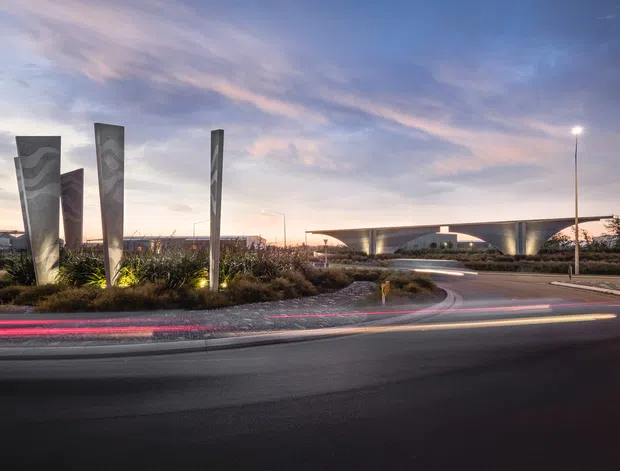 A dusk exterior view of a landscaped civic or commercial precinct, with tall sculptural metal panels in a planted traffic island, curving roads, street lighting, and a low sweeping modern building with an arched or domed central form in the background. Vehicle light trails emphasize the active roadway and contemporary public-realm setting.