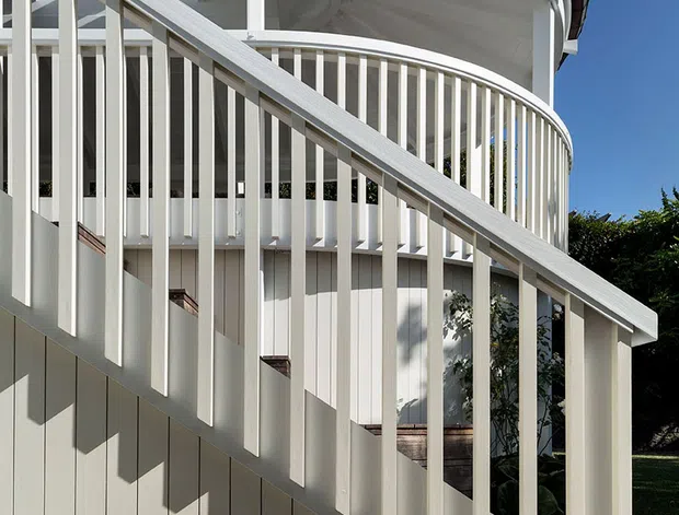 Exterior architectural detail of a white, curved building featuring a sweeping circular balcony or veranda, prominent white vertical balustrades, a diagonal stair railing, and a distinctive radial ribbed soffit beneath a dark curved roof edge. Strong sunlight creates crisp shadows across the pale cladding and textured concrete base, set against a clear blue sky and greenery.