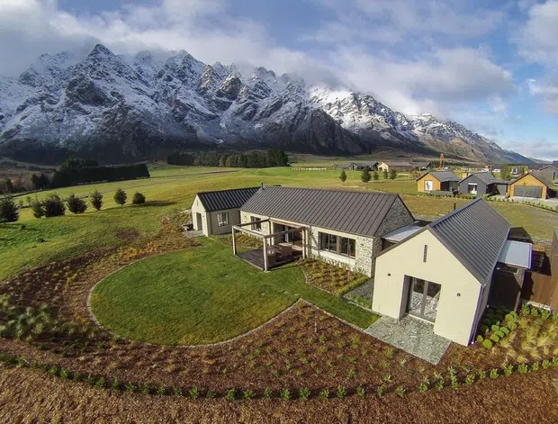 Aerial view of a contemporary rural residential property set among cultivated fields, with several grey metal-roofed buildings, landscaped gardens, and dramatic snow-capped mountain ranges in the background.
