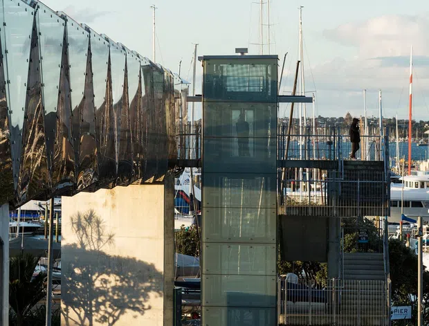 Exterior architectural photograph of a contemporary waterfront building beside a marina. The structure features a tall glazed lift or circulation tower, dark metal railings, exposed structural elements, and a highly reflective angular metal-clad wing. Boats, masts and harbour infrastructure are visible behind it, with a multilane road in the foreground.