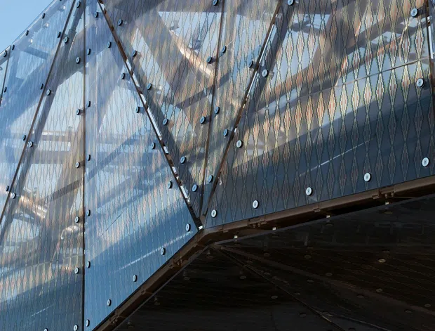 Close-up upward-looking view of a contemporary architectural facade and projecting canopy. The building features extensive transparent glass panels fixed with exposed circular fasteners, angular steel framing, patterned translucent or reflective glazing, and a dark ribbed metal soffit beneath the overhang. The image emphasizes geometric structure, transparency, and modern construction detailing against a clear blue sky.