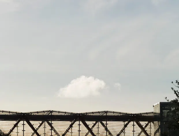 A backlit exterior view of a large enclosed steel truss bridge or elevated pedestrian structure spanning the frame, with visible diagonal bracing, mesh-like side panels, and a dark glazed building connection at the right. Trees and a distant wooded ridge sit below a pale, cloud-filled sky.