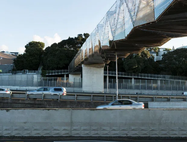 Exterior architectural photograph of a large elevated pedestrian bridge or covered walkway spanning above a multilane roadway. The structure has extensive translucent or glass-like cladding, exposed dark steel framing, and substantial concrete support columns. Urban residential buildings and trees are visible in the background, while traffic and road markings occupy the foreground.