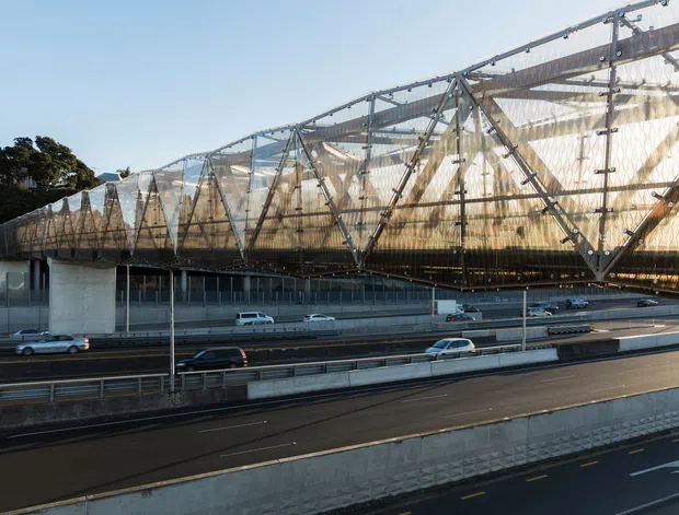 Exterior architectural view of a large elevated transport or infrastructure structure spanning a multi-lane roadway. The structure features a long transparent and translucent enclosure with exposed steel trusses, diagonal bracing, and substantial concrete supports. Multiple lanes of traffic run beneath and alongside it, with trees and open sky in the background.
