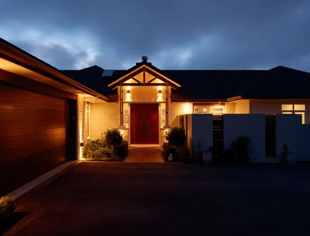 Twilight exterior view of a single-storey residential home arranged around a driveway or forecourt. The facade combines dark tiled pitched roofs, warm illuminated timber-look wall cladding, light masonry or rendered panels, a prominent gabled entry porch with exposed timber framing, a red front door, and an integrated garage on the left. Low planting and privacy screens frame the entrance.