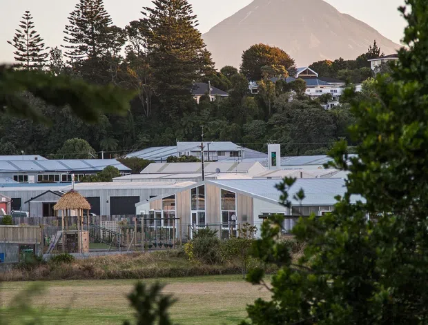 Exterior landscape view of a low-rise contemporary building complex with pale metal roofs and extensive glazing, seen across a grassy open area and partially framed by trees. A prominent steep volcanic mountain rises behind the suburban settlement, creating a strong mountain setting. The image is photographed from outdoors and is dominated by the building exterior and surrounding landscape.