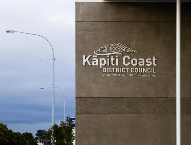 Exterior view of the Kapiti Coast District Council civic building, featuring a large textured concrete or rendered facade panel with prominent council signage, a broad projecting roof canopy, and a white structural column. The streetscape, curved streetlights, paved forecourt, planted verge, and overcast coastal-looking sky are visible around the building.