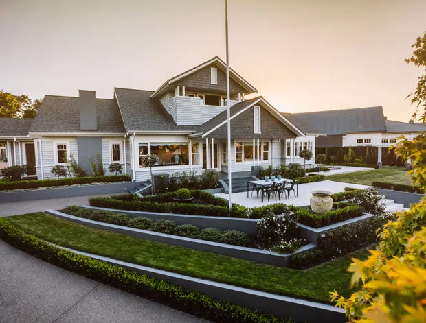 A large, multi-wing suburban residence with a pitched gable roof, white weatherboard-style exterior, dark grey roofing, covered verandas, and prominent white-framed windows. The home is surrounded by formal tiered garden beds, clipped hedges, paved paths, and a central outdoor dining area, viewed from the landscaped front approach in warm evening light.