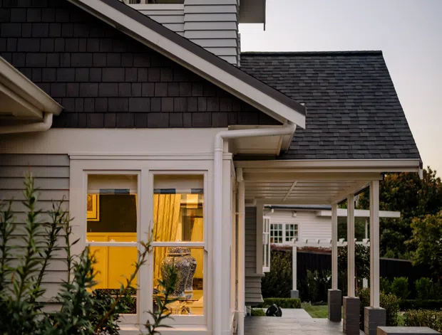 Photograph of a refined residential exterior showing a multi-level home with dark shingle-clad gabled roofs, pale horizontal weatherboard siding, white trims, and a covered entry walkway. A small upper balcony is visible above the entry, while landscaped planting borders dark tiled steps with integrated warm lighting. The scene has a traditional coastal-suburban character.