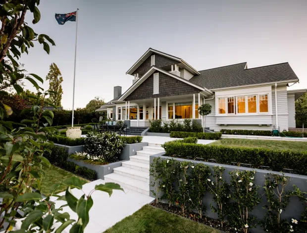 Exterior view of a large, carefully landscaped suburban house with white weatherboard cladding, dark shingle-style pitched roofs, prominent gables, covered verandah, and warm-lit windows. Formal hedges, terraced garden beds, clipped topiary, and a central stairway lead up to the entrance. A flagpole stands in the front garden.