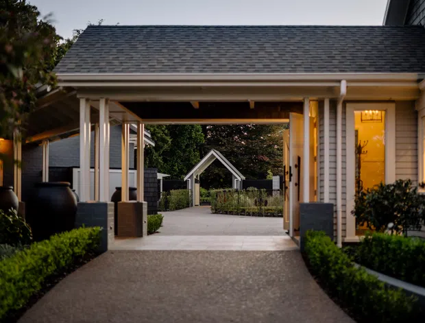 A dusk exterior view of a refined residential entry court or covered passage. A broad gabled roof canopy connects light-coloured weatherboard-clad buildings, with white posts and trim, landscaped hedges, large dark planters, and a paved walkway leading toward a small gabled structure in the garden. Warm interior lighting glows through the windows and doorway, creating an inviting, formal atmosphere.