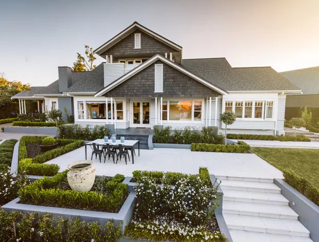 Exterior view of a large, symmetrical suburban house with white weatherboard cladding, grey shingle-style gabled roofs, prominent dormer-like gable forms, and white-trimmed windows. The property features a formal landscaped garden with geometric hedges, paved terraces, outdoor dining furniture, and broad steps leading to the home.