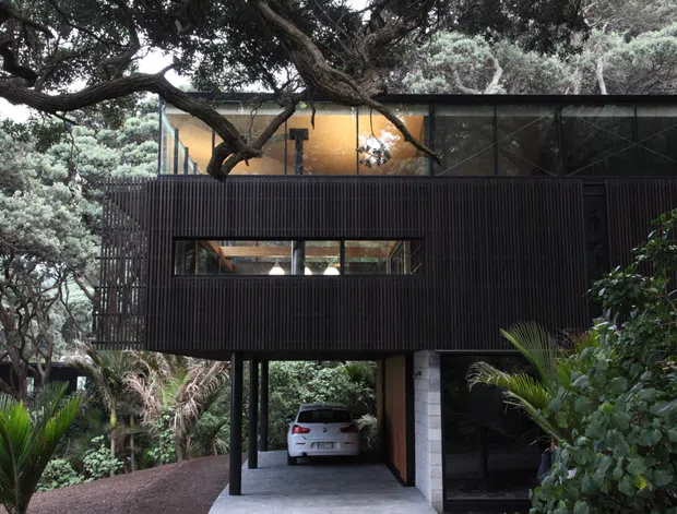 Exterior view of a dark, elevated modern house nestled among dense mature vegetation. The building has a rectilinear form, extensive dark vertical ribbed cladding, large glazed upper-level openings, and a covered carport beneath the raised living volume. A substantial tree canopy crosses in front of the facade, reinforcing the integrated bush setting.