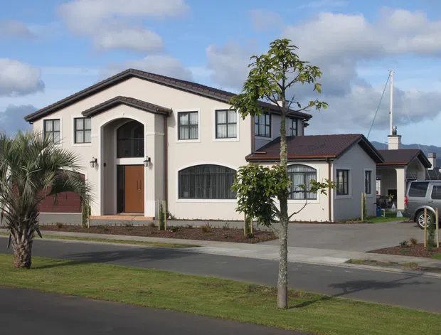 Exterior street view of a multi-level suburban house with cream rendered walls, dark tiled gabled roofs, an arched recessed entry, timber front door, and dark-framed windows. A connected lower wing and driveway are visible, with landscaped street planting and marina masts in the background.