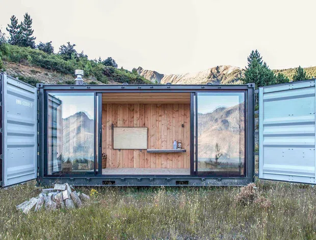 A compact container-style cabin or retreat set in a mountainous alpine landscape. The open dark metal-clad shell frames a timber-lined interior with large glazed openings, a small central shelf and wall-mounted panel, while the surrounding mountain scenery is reflected in the glass. The image emphasizes the prefabricated, transportable structure and its integration into the rugged natural setting.