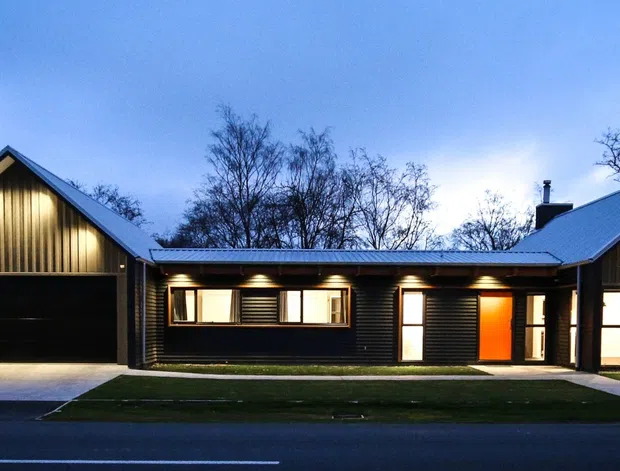 Twilight exterior view of a low-profile contemporary home composed of two steep gabled wings connected by a recessed central section. The facade features dark vertical cladding, warm timber-toned gable detailing, large garage doors, illuminated windows, and a prominent orange entry door, with a simple lawn and driveway in front.